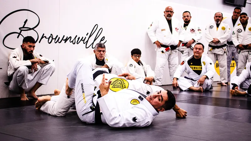 Adult students in white gis watch from the edge of the mat while an instructor demonstrates a ground technique