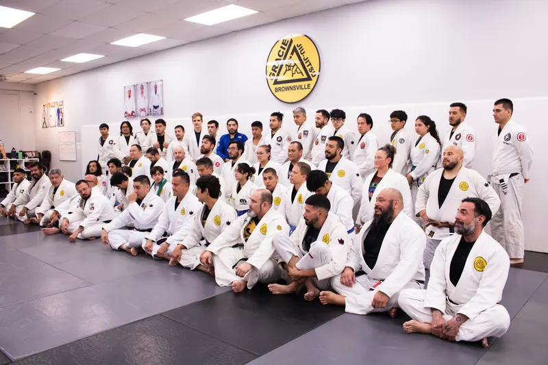 A large group of jiu-jitsu students in white and blue gis seated in rows on the mat under the Gracie Jiu-Jitsu Brownsville crest at the BJJ Spring Seminar