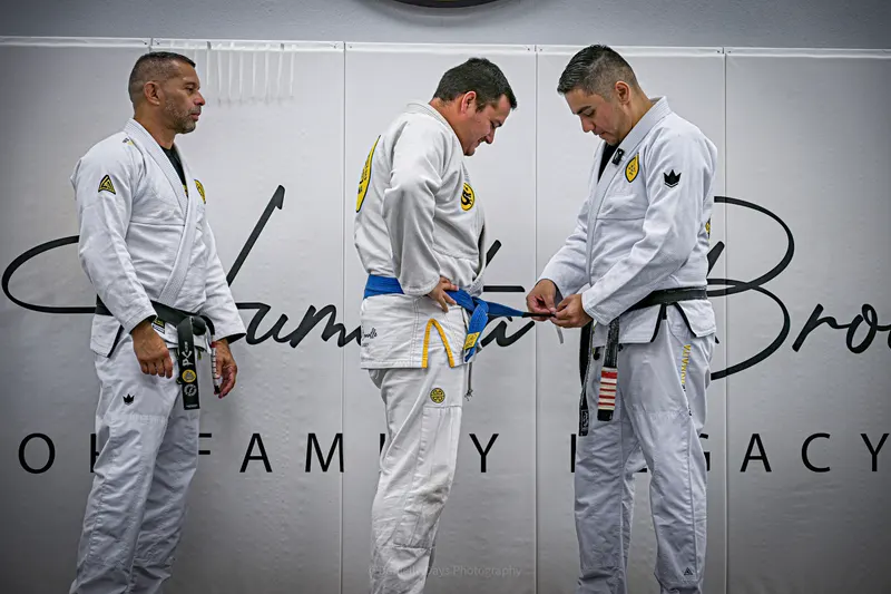 A black belt ties a new blue belt on a student while another black belt looks on, in front of the Gracie Humaita Brownsville Honor Family Legacy wall