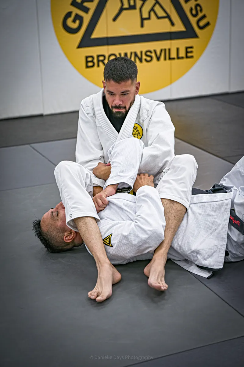 An instructor in a white gi demonstrates a ground technique on a partner in front of the Gracie Jiu-Jitsu Brownsville crest