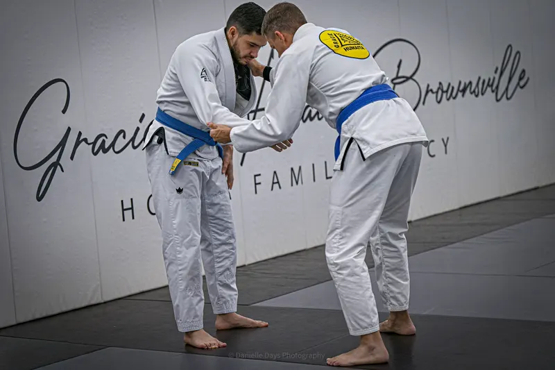 Two blue belts grip each other's gis during a standing drill in front of the Gracie Humaita Brownsville Honor Family Legacy wall