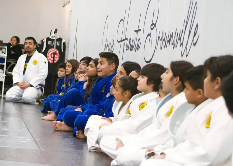 Children in blue and white jiu-jitsu gis sitting in a row along the wall listening to their instructor during a kids class